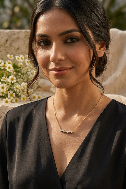 A woman with dark hair wearing a delicate gold necklace with a small cluster of clear stones, photographed in warm natural light against a soft beige stone background with white flowers.